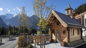 A wooden cabin with a bell tower, surrounded by a paved area with benches and a small garden. - Stylish Alpine Chalet With Carport (Neukirchen am Grossvenediger)