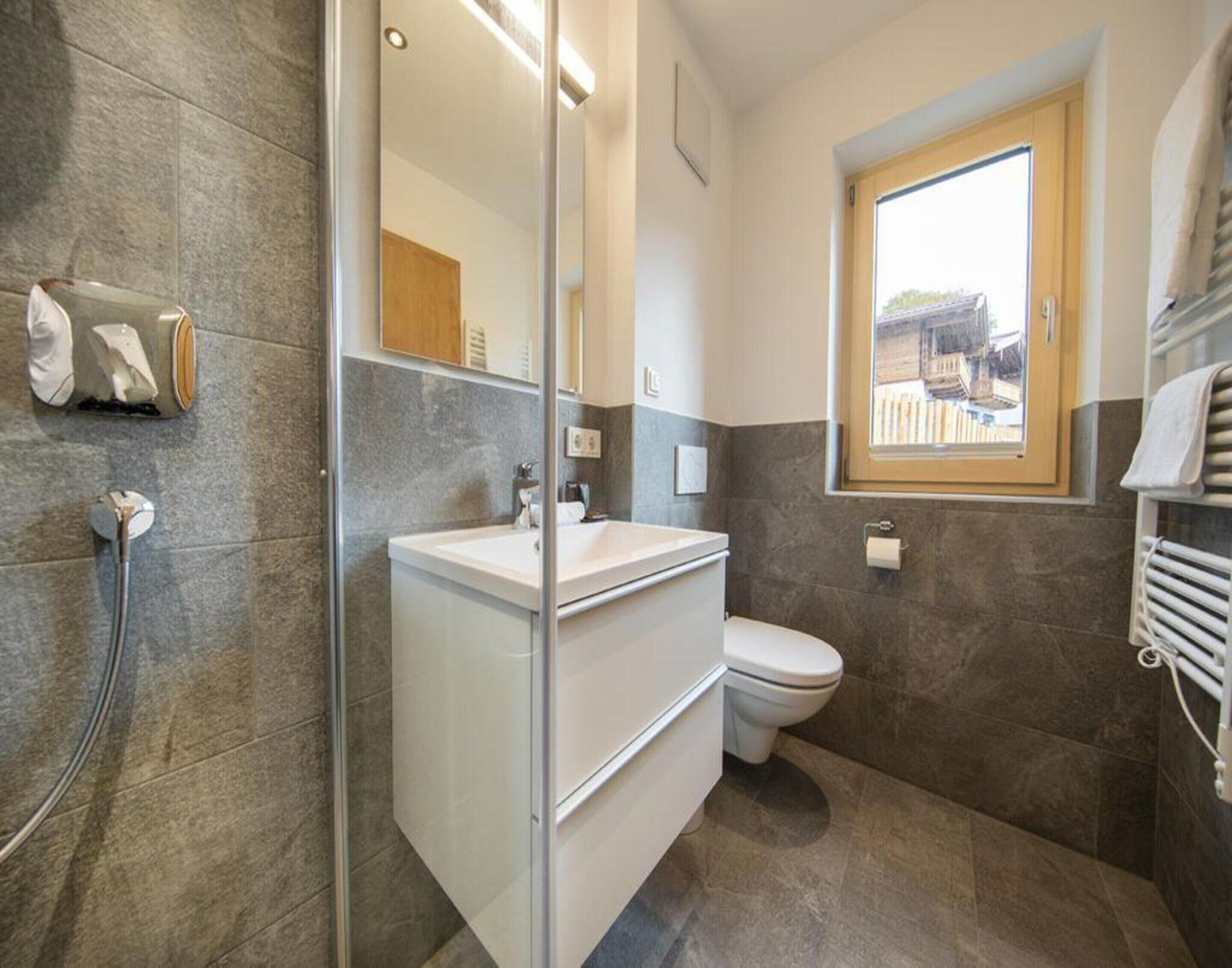 A modern bathroom with a glass shower enclosure, a white sink, and a toilet. There is a window providing natural light and a view of the outside.