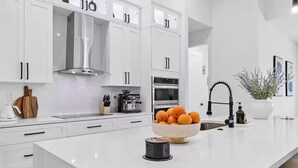 A modern kitchen with white cabinetry, a stainless steel range hood, a coffee machine, and a bowl of oranges on the counter. - Luxury Waterfront Retreat! Hot Tub and Heated Pool. (Cape Coral)