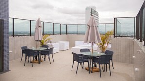 Eine Dachterrasse mit einem Glastisch und Stühlen, umgeben von weißen Sofas und zwei Sonnenschirmen.