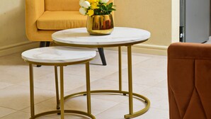 Una habitación con un sillón amarillo, dos mesitas redondas con patas doradas y un florero sobre una de ellas.