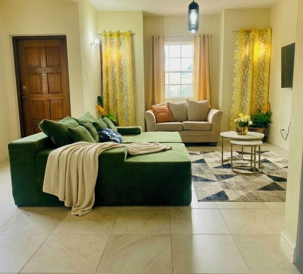 Un salon avec un canapé vert, un canapé beige, une petite table ronde et un tapis à motifs.