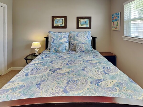 Een slaapkamer met een bed, twee afbeeldingen in lijsten aan de muur, een nachtkastje met een lamp en een raam met jaloezieën.