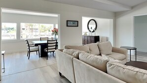 Ein modernes Wohnzimmer mit einem beigefarbenen Ecksofa, einem Esstisch mit Stühlen und einem kleinen Beistelltisch. Ein großes Fenster lässt viel Tageslicht herein und bietet einen Blick ins Freie.