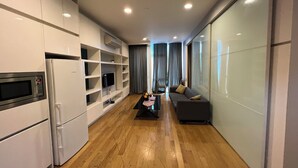 Cozinha moderna com eletrodomésticos embutidos, área de refeições com sofá e mesa de centro, e uma janela grande com cortinas.