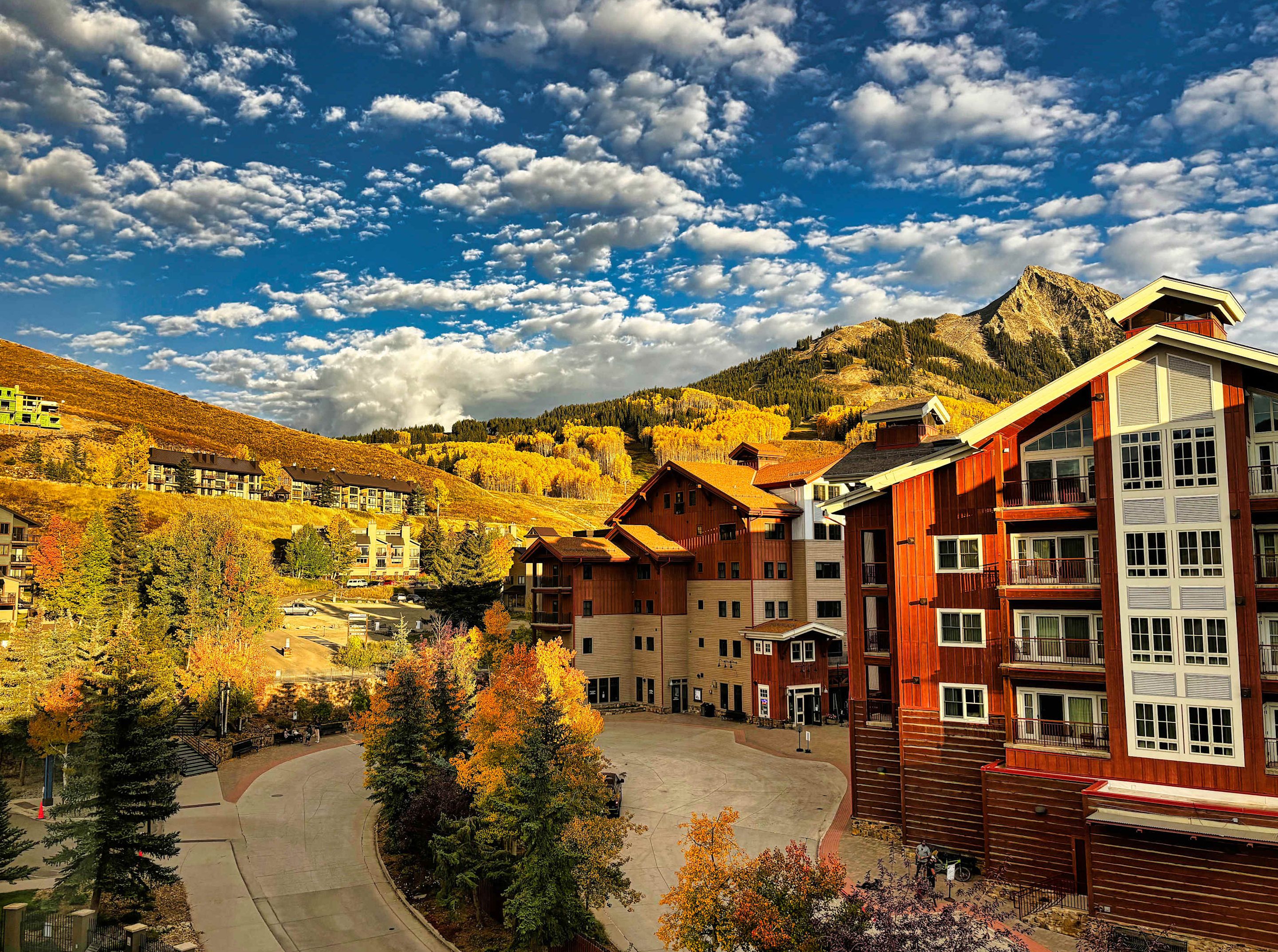 A mountain resort with multi-story buildings, a central courtyard, and a backdrop of autumn-colored trees and a mountain.