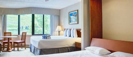 A hotel room with a bed, a desk, a chair, a ceiling fan, and a window with a view of greenery.
