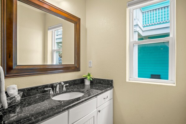 A bathroom with a black marble sink, a large mirror, and a window with a view of a blue house. - New Listing! Gulf Views. (Port Aransas)