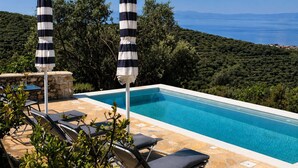 A rectangular infinity pool with a clear blue edge, surrounded by lounge chairs and striped umbrellas, overlooking a mountainous landscape. - Niki's Tower - Exclusive Serene Seaview Stone Gem (Kalamata)