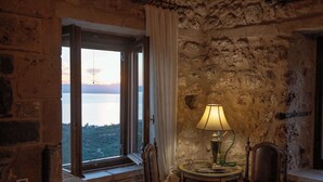 A room with a stone wall, a window with curtains, a table with a lamp, and a chair. - Niki's Tower - Exclusive Serene Seaview Stone Gem (Kalamata)