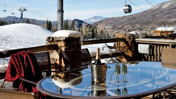 A round glass table with chairs, a bottle, and glasses on a wooden deck with a stone wall and a ski lift in the background. - 700 Ute Apartment #703 4 Bedroom Home (Aspen)
