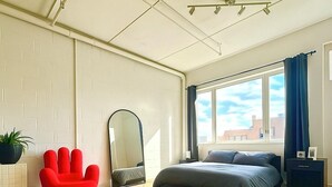A modern bedroom with a large bed, a red chair, a mirror, and a view of the city through the window.