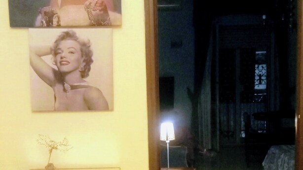 A room with a marble floor, a door leading to another room, a small table with a lamp, and two framed pictures on the wall.