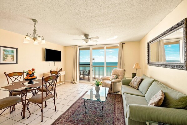 A living room with a dining area, a sofa, a TV, and a balcony with a view of the ocean. - Pelican Beach 0602 1 Bedroom Condo (Destin)