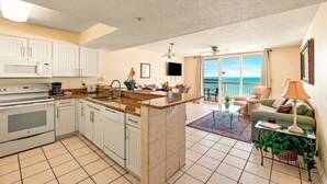 A spacious kitchen with a central island, a dining area with a view, and a living room with a sofa and a TV. - Pelican Beach 0602 1 Bedroom Condo (Destin)