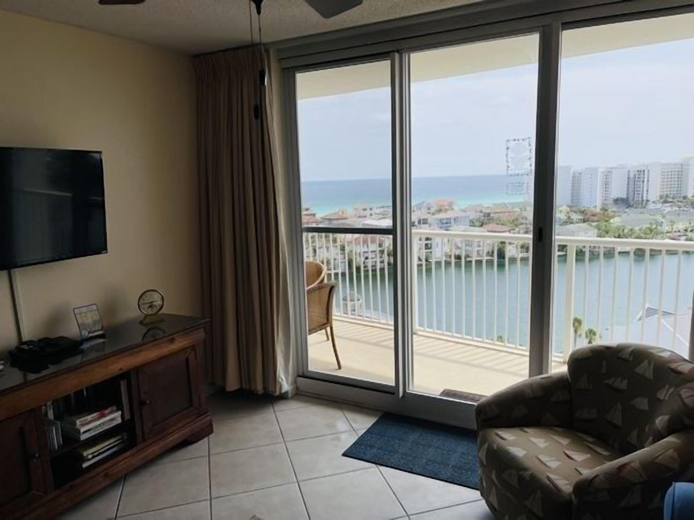 A room with a TV, a wooden cabinet, a patterned armchair, and a large sliding glass door leading to a balcony with a view of the sea and buildings.