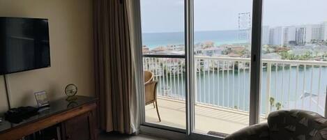 A room with a TV, a wooden cabinet, a patterned armchair, and a large sliding glass door leading to a balcony with a view of the sea and buildings.