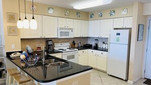 A kitchen with white cabinets, a black countertop, a stainless steel sink, a microwave, a stove, a refrigerator, and three wooden bar stools.