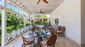 A covered outdoor dining area with a round table, chairs, ceiling fans, and a view of lush greenery. - Rosedale 3 Home (Worthing)