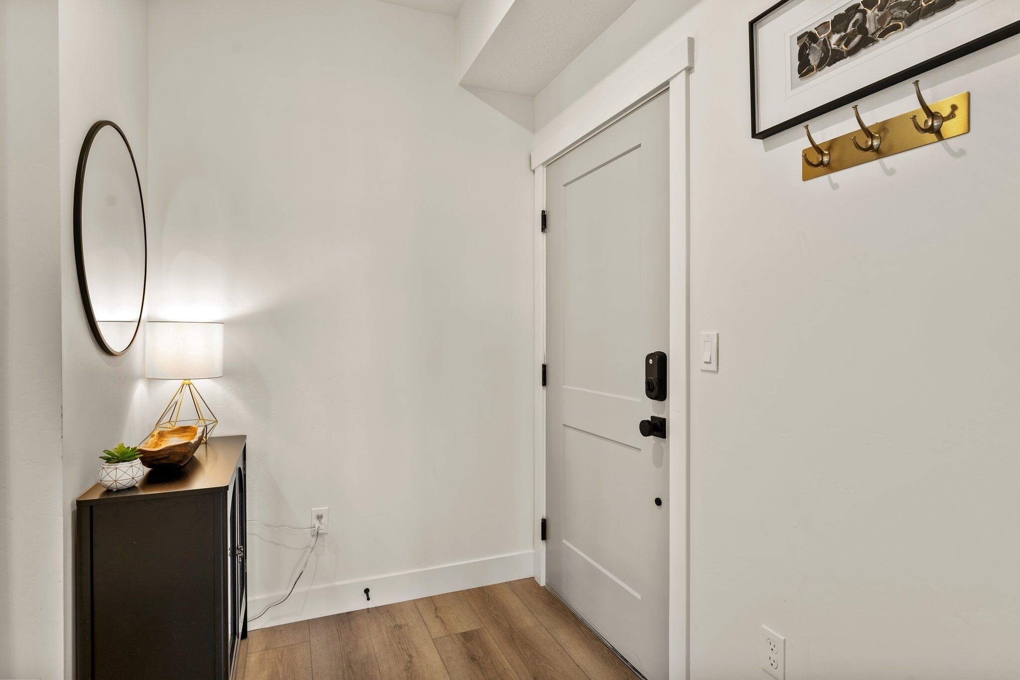 A hallway with a closed door, a wall-mounted lamp, a framed artwork, and a small wooden cabinet with a plant.