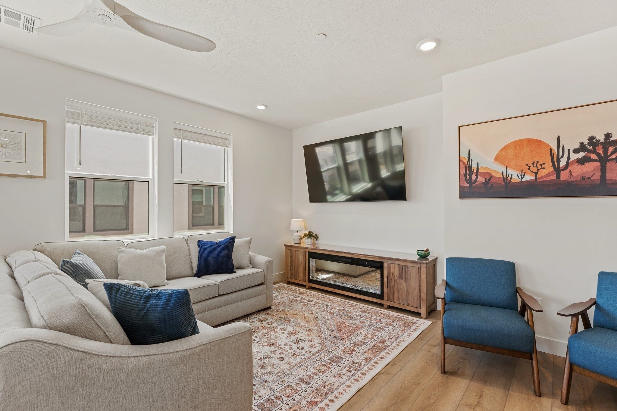 A living room with a beige sofa, blue armchairs, a patterned rug, a wooden TV stand, and a desert-themed painting.