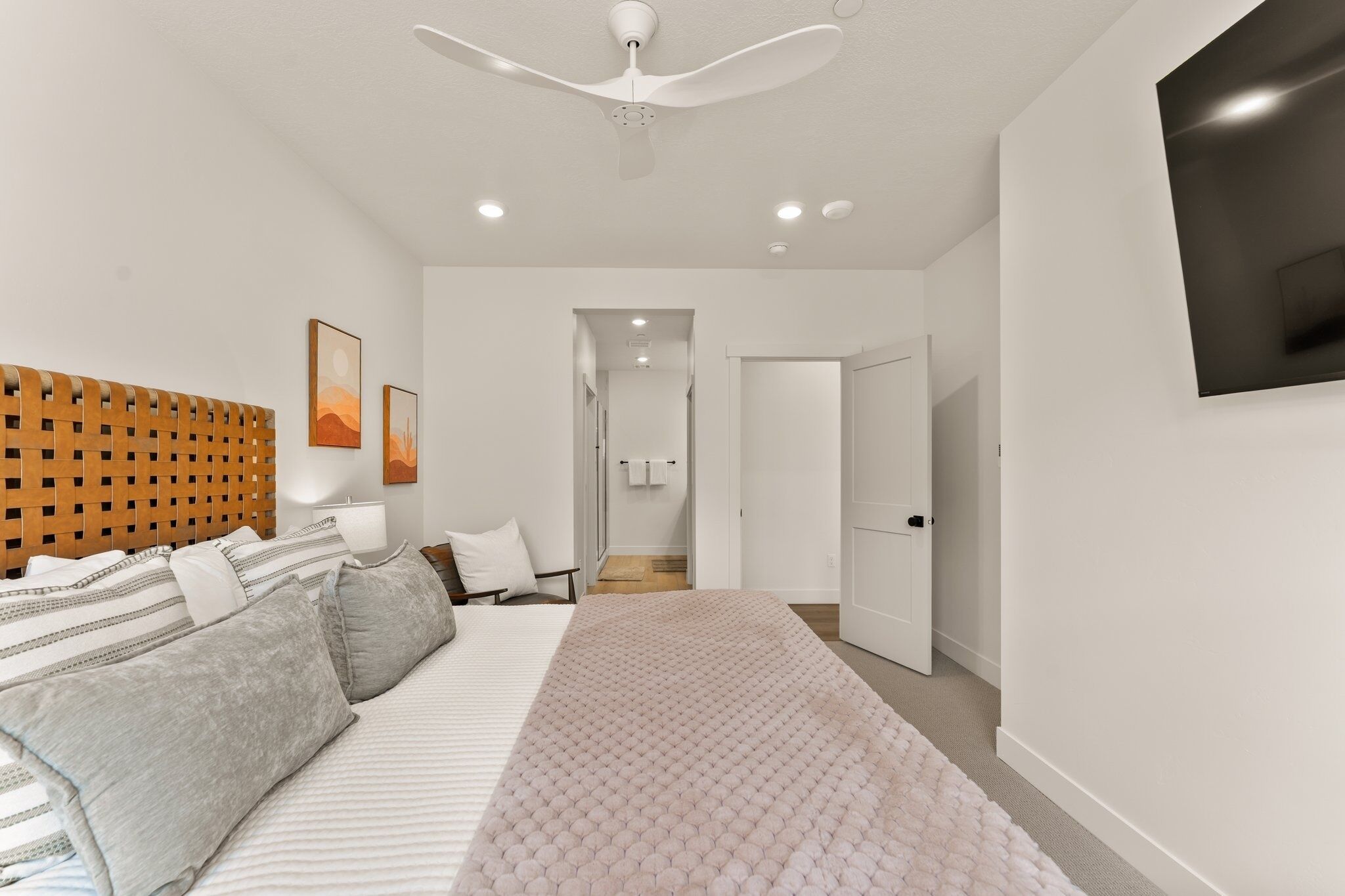 A modern bedroom with a bed, a ceiling fan, a TV mounted on the wall, and a hallway leading to a bathroom.