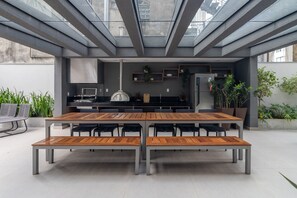 Ein moderner Essbereich im Freien mit einem langen Holztisch und Metallstühlen, einer Küchenzeile mit Kühlschrank und Schränken sowie Zimmerpflanzen.