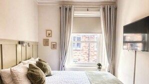 Una cama bien hecha con almohadas, una mesita de noche, un televisor y una ventana con cortinas.