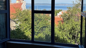 A window view of a coastal cityscape with buildings, greenery, and a clear sky. - Dafne Konukevi (Bozcaada)