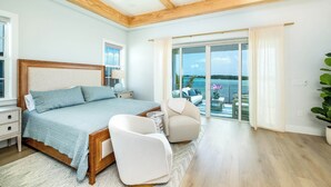 Een slaapkamer met een bed, nachtkastjes, een stoel, een balkon met uitzicht op zee en een plafondventilator.