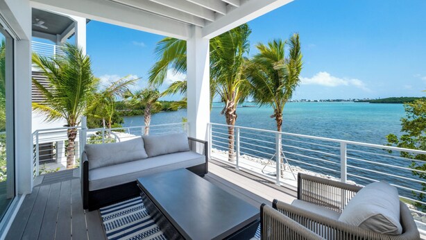 Een moderne buitenruimte met een bank en een tafel, met uitzicht op een heldere blauwe zee met palmbomen.