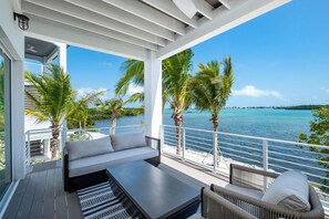 A modern outdoor seating area with a sofa and a table, overlooking a clear blue sea with palm trees. - Brand New Oceanfront 5BR - Luxury Stay w/ Elevator & Boat Slip Included (Marathon)