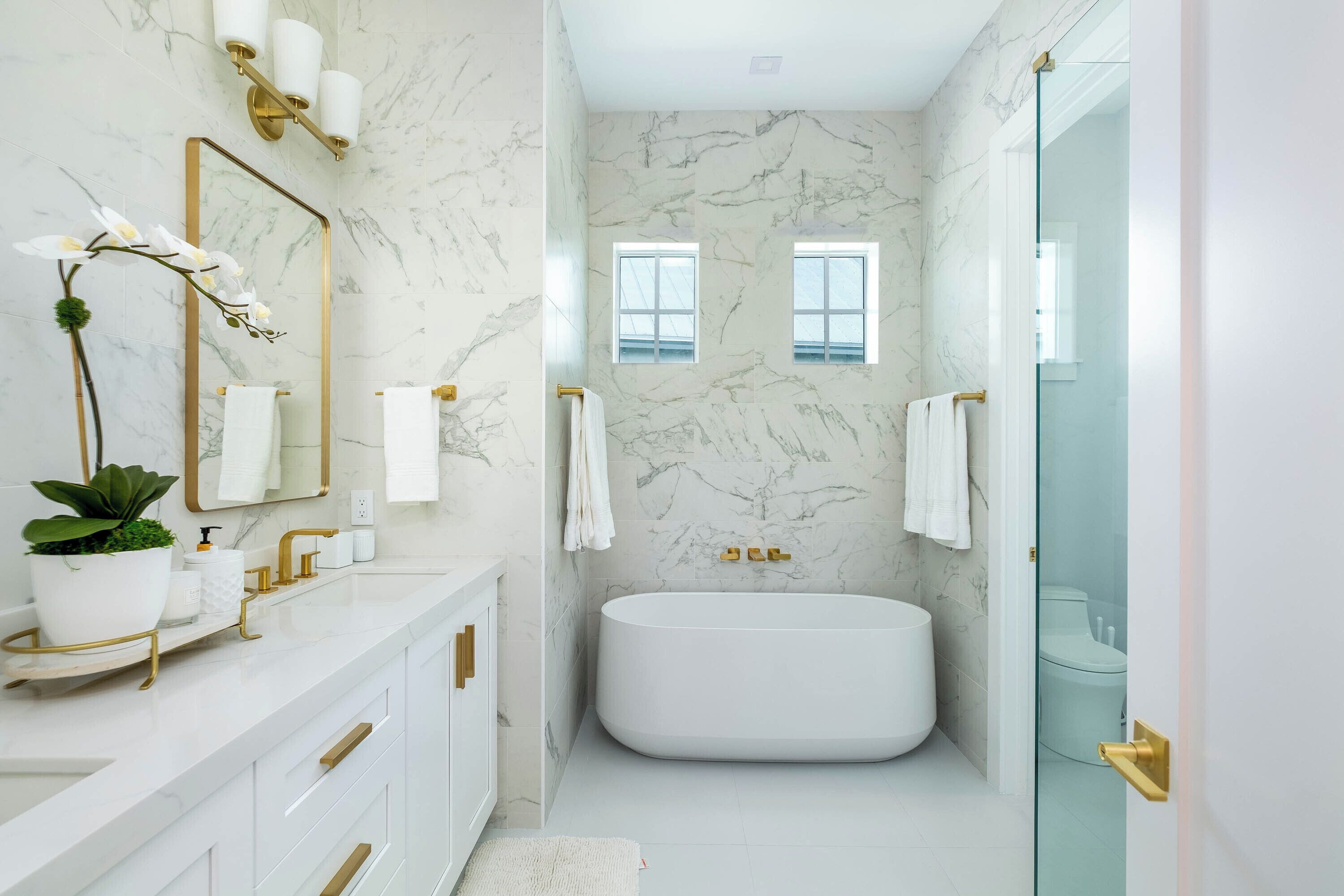 A modern bathroom with a marble wall, a freestanding bathtub, and a vanity with gold fixtures.