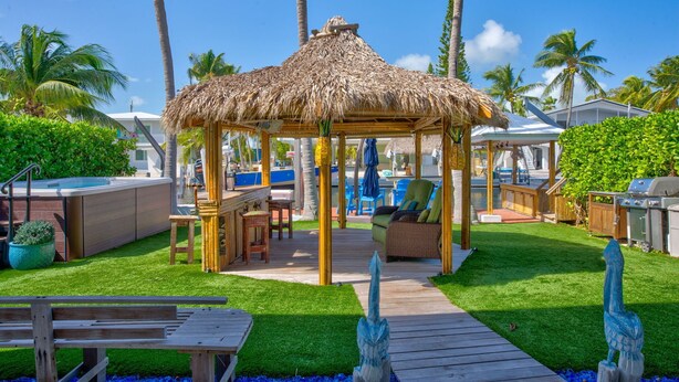 A thatched gazebo with a pool and outdoor furniture, surrounded by palm trees and a well-maintained lawn.