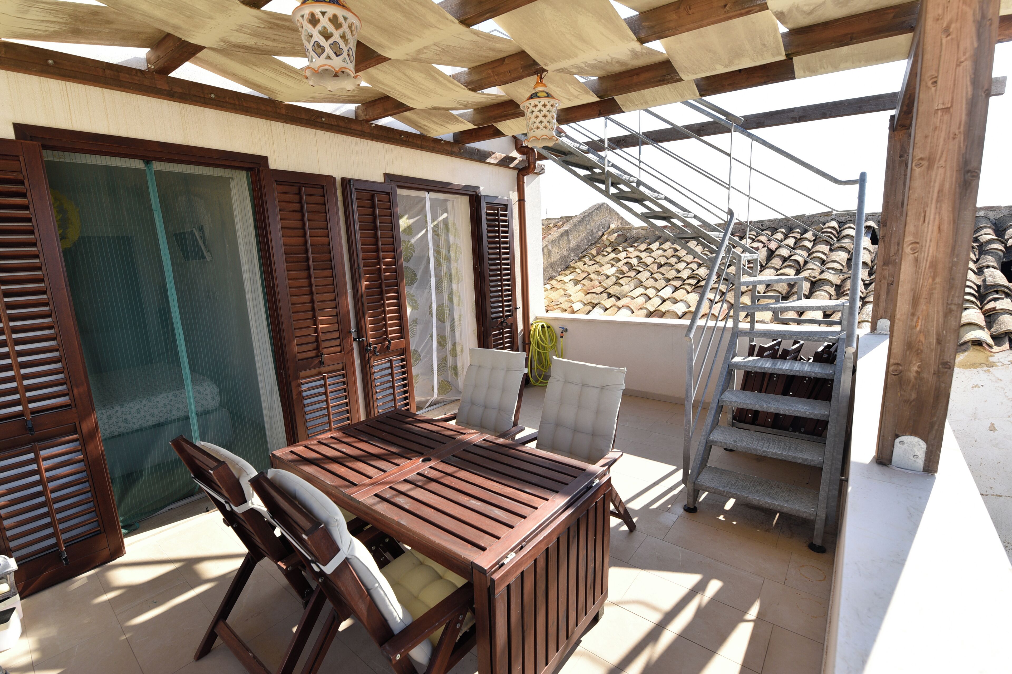 A covered outdoor patio with a wooden table and chairs, a metal chairlift, and a view of a tiled roof in the background.