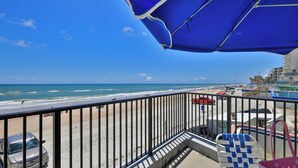 A balcony with a view of the beach, a red car, and beach umbrellas.