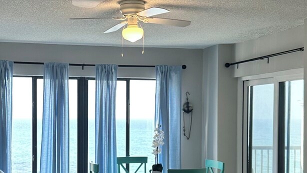 A living room with a large window, a ceiling fan, and a dining area with chairs and a table.
