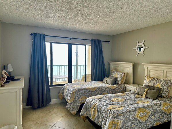 Habitación de hotel con dos camas, un escritorio, una silla y vista al mar a través de amplias ventanas.