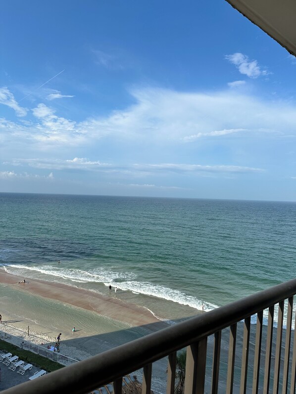 Vista a la playa desde un balcón con barandilla, océano y cielo despejado.