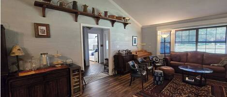 Amplia sala de estar con piso de madera, sofá de cuero marrón, una mesa de centro y una chimenea. Cuenta con un ventilador de techo y una ventana con persianas.