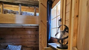 Een houten blokhut interieur met een stapelbed, een klein bureau met een ventilator en een raam met gordijn.