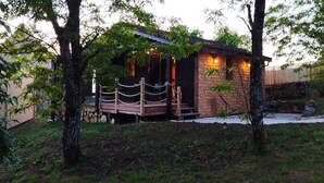 Een rustieke chalet met een houten terras en stenen muren, omgeven door bomen en een grindpad.