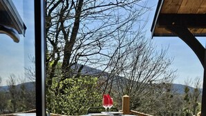 Een houten terras met een tafel, stoelen, een touwbordes en uitzicht op bomen en heuvels.