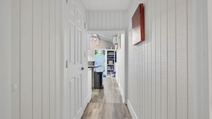 A narrow hallway with white paneling and a wooden floor, leading to a room with a blue door and a white wall-mounted shelf.