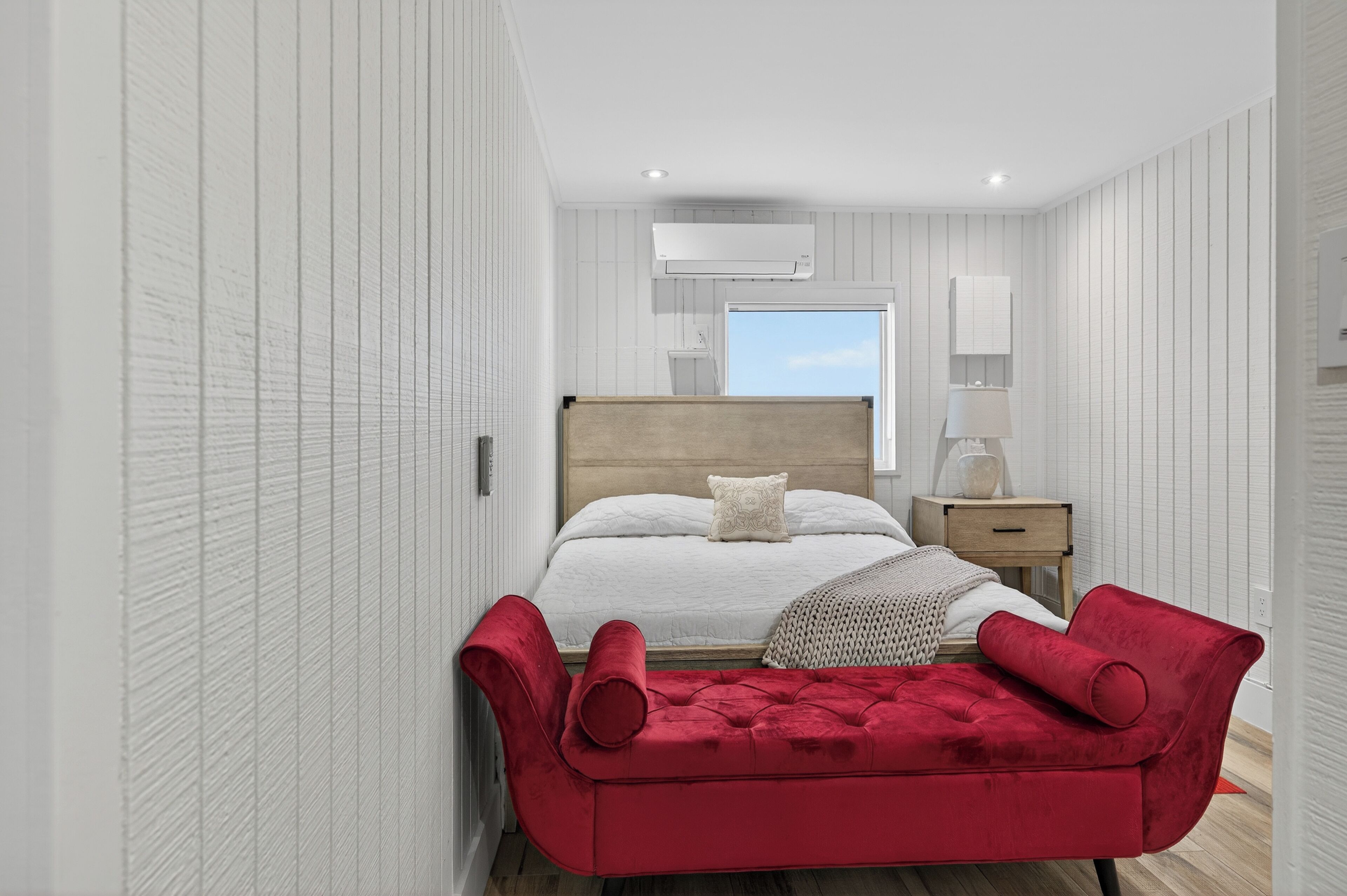 A bedroom with a large bed, a wooden headboard, a red upholstered bench, a nightstand, and a window with a view of the sky.