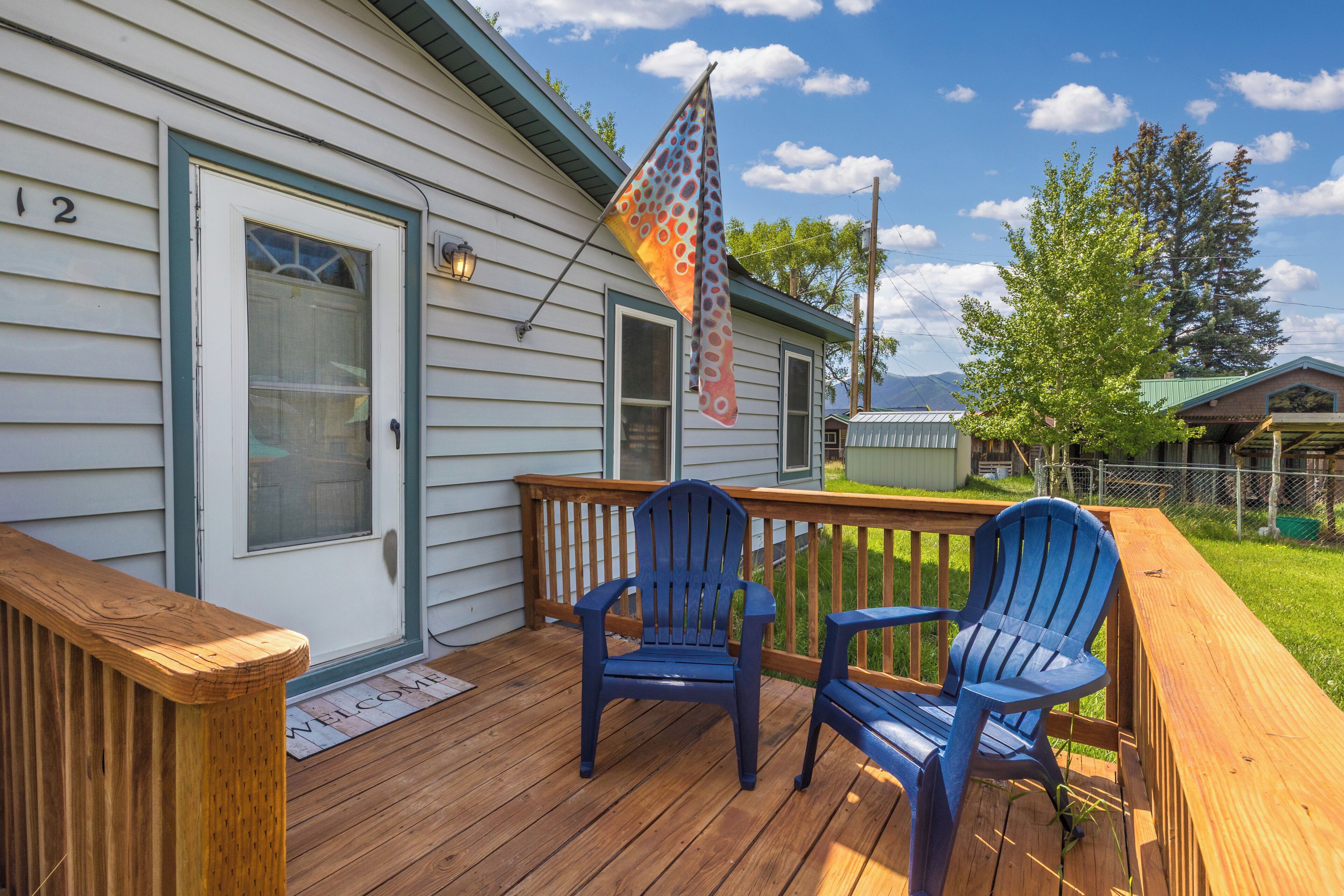 A wooden deck with two blue Adirondack chairs, a flag, and a house with a number '12' on the door.