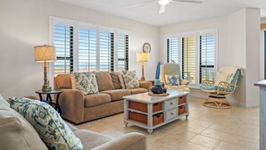 A living room with a beige sofa, a blue armchair, a white coffee table, and a ceiling fan. - Oceanfront 1st Floor Walk-Out w/ Ocean Views, Steps to Beach & Pool - 4BR 3BA (Indian Beach)