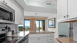 A modern kitchen with white cabinetry, a granite countertop, a stainless steel refrigerator, and a built-in microwave. - Oceanfront 1st Floor Walk-Out w/ Ocean Views, Steps to Beach & Pool - 4BR 3BA (Indian Beach)