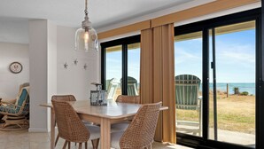 A dining area with a wooden table and wicker chairs, a hanging light fixture, and a view of the ocean through large sliding glass doors. - Oceanfront 1st Floor Walk-Out w/ Ocean Views, Steps to Beach & Pool - 4BR 3BA (Indian Beach)
