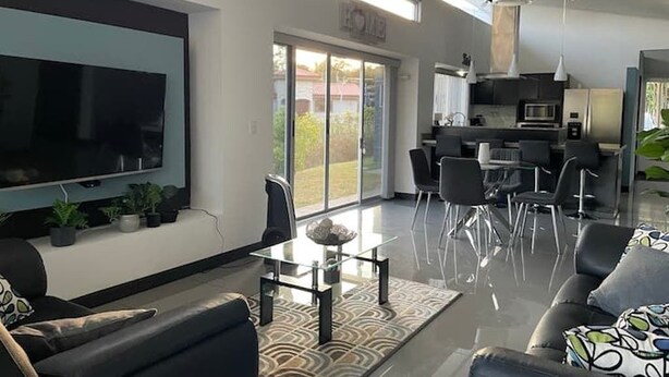 Un saló modern amb televisió de pantalla plana, taula de centre de vidre i sofà. Es veu una zona de cuina amb una taula de dinar i cadires a través d'una porta de vidre.